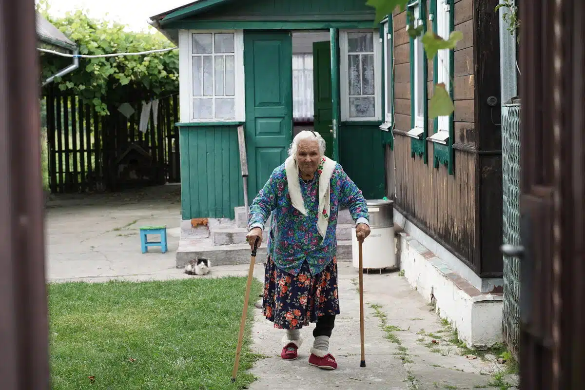 An older women walks with the support of 2 canes, one in each hand, looking directly at the camera as she takes a step. 