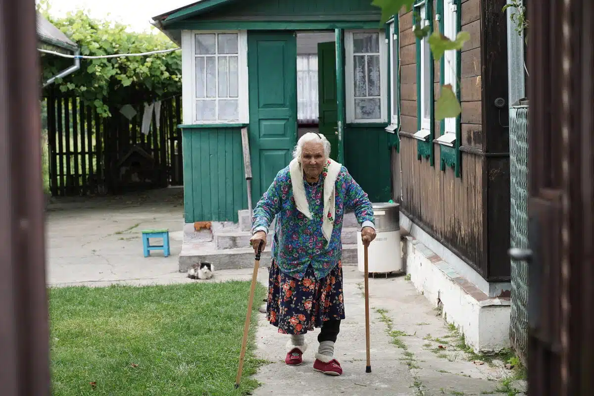 An older women walks with the support of 2 canes, one in each hand, looking directly at the camera as she takes a step.