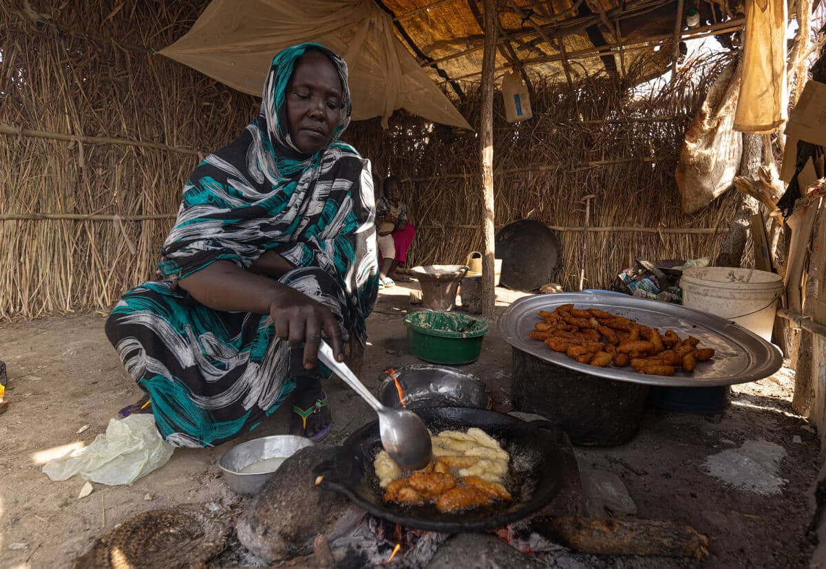 A woman sits on the floor of her shelter, cooking food and spooning it into a plate. 