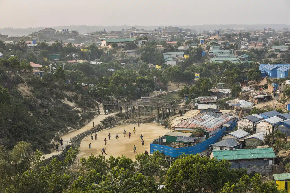 bird's-eye-view picture of children playing soccer on a dirt field surrrounded by vegetation on the left and a refugee camp on the right