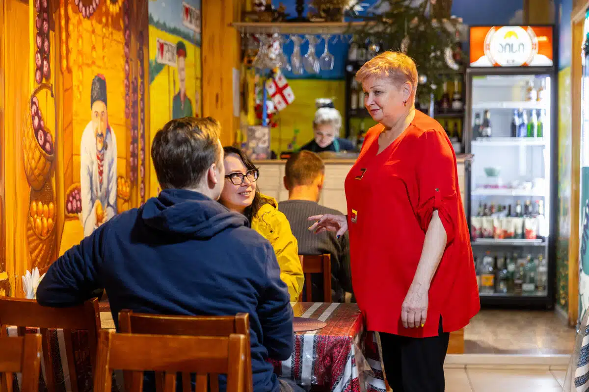 A woman talks to two customers in a cafe sitting across from each other at a table