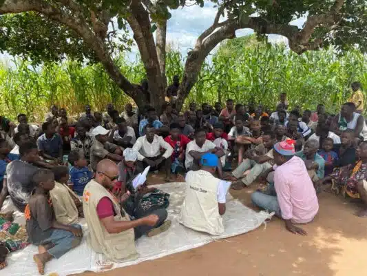 A group of people sit on the dirt ground under a tree