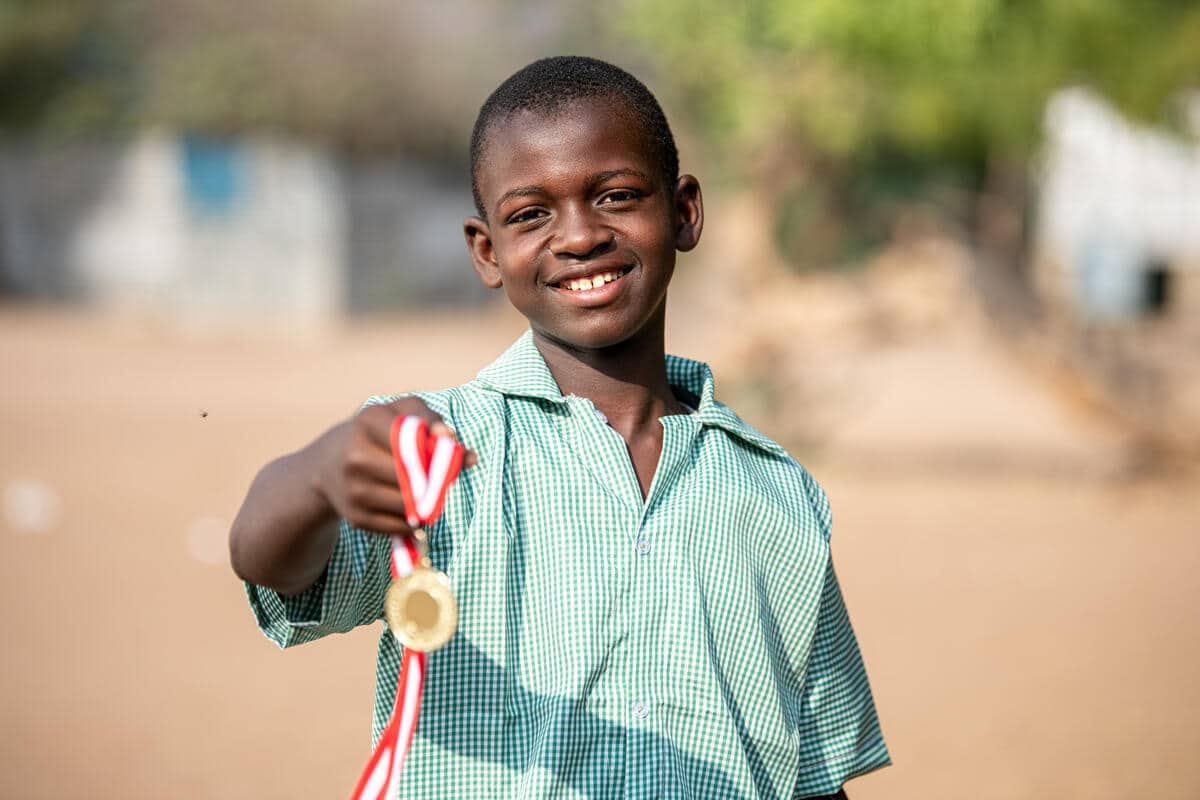 A close-up of a young boy smiling and holding out a gold medal
