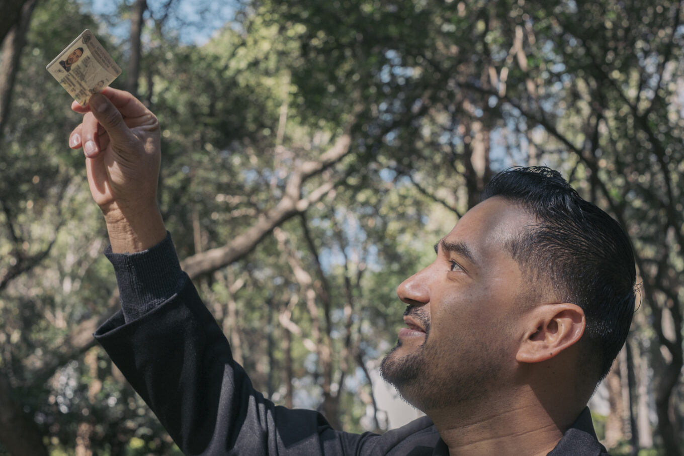 Man in front of a forest background holding up a Mexican ID Card.