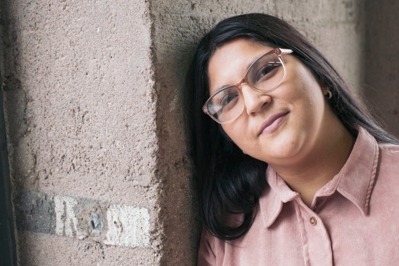 Woman with glasses in pink button up shirt leaning against a cement wall.