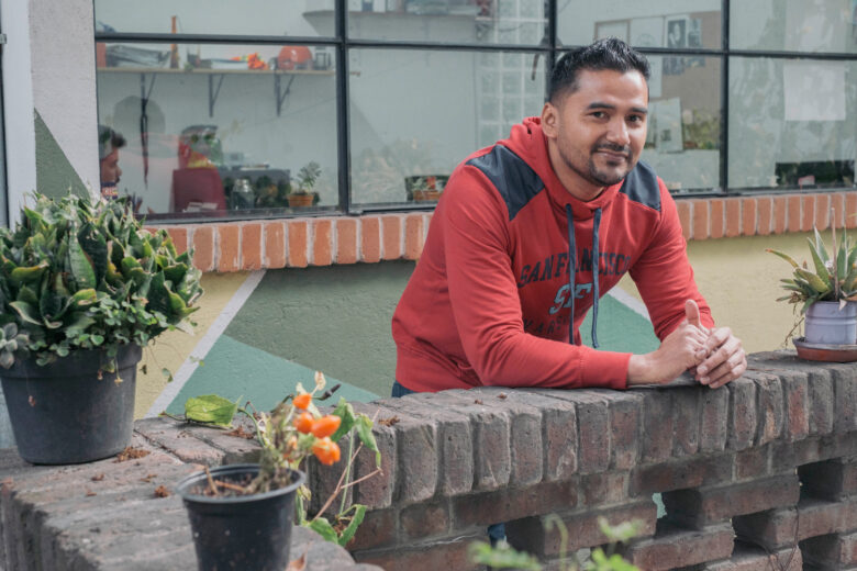 Man with red hoodie leaning against a stone fence. He is in front of windows. There are potted plants next to him on the fence.