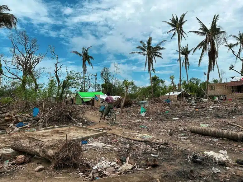 Village has been ;eft destroyed by a cyclone. There is debris and a man biking through the devestation.