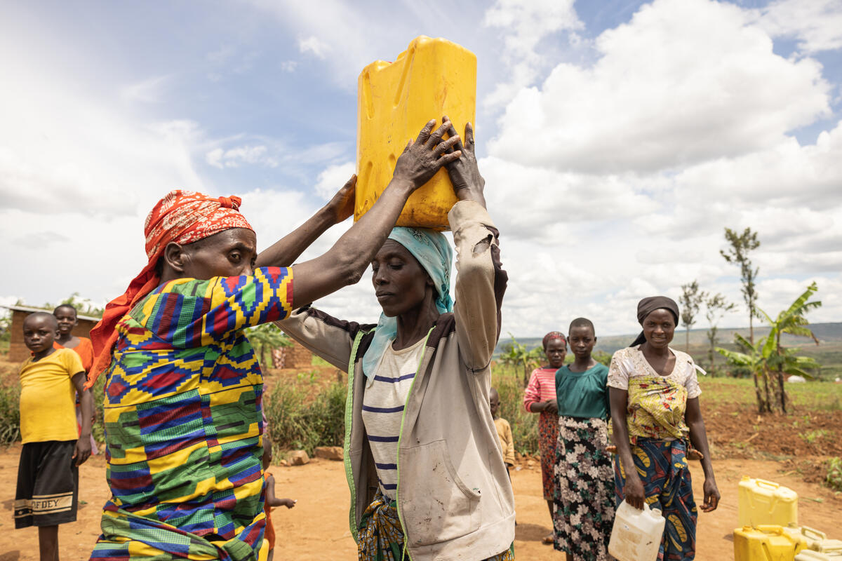 In the foreground, an older woman in a colourful dress and red headscarf helps hoist a bright yellow plastic jar filled with water on the head of a younger woman dressed in a fraying grey hoody and white t-shirt with black horizontal stripes. In the background a group of women and children in Nakivale refugee settlement, Uganda, wait for their turn to collect water.© UNHCR/Esther Ruth Mbabazi