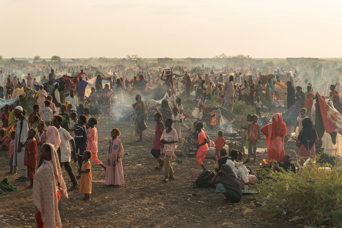 Thousands of Sudanese and South Sudanese refugees gather in a dusty field with camp fire smoke rising over the plain at the Joda border crossing in Sudan's White Nile State in November 2023.