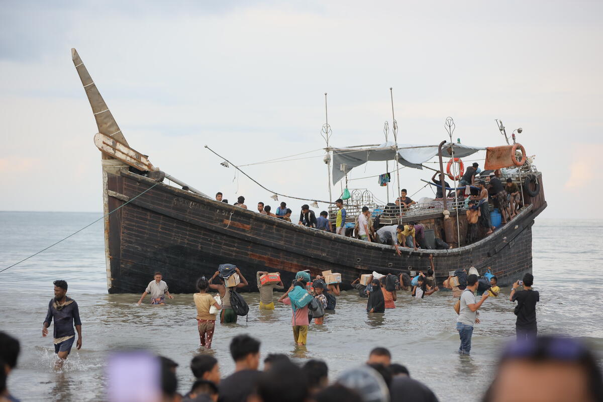 In the middle of the picture, a wooden boat in the water full of refugees. Many people are walking towards the boat carrying boxes and bags in the water.