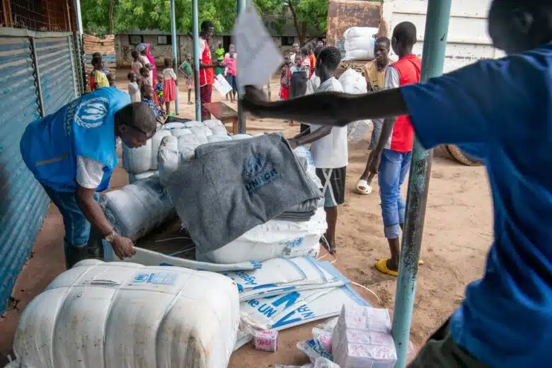 Man in blue UNHCR vest leans over a pile of UNHCR branded emergency items.