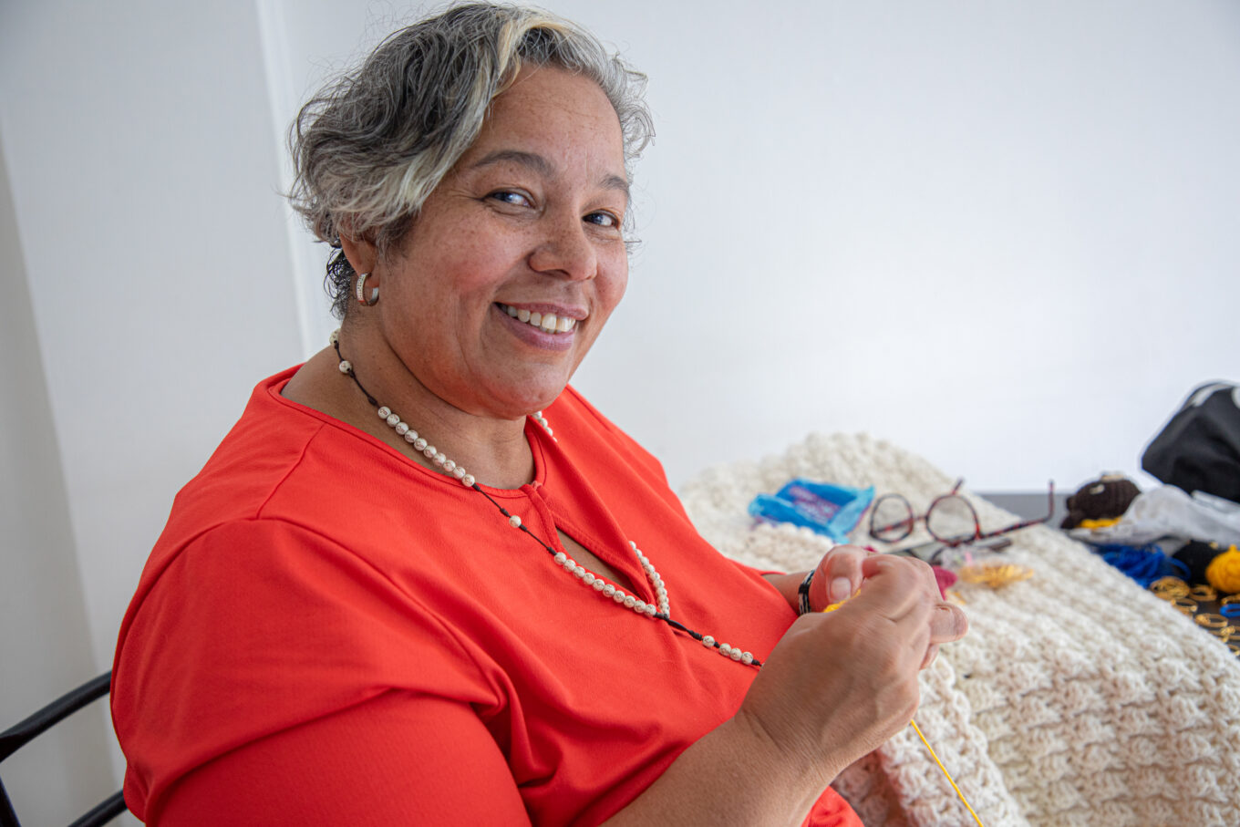 Woman wearing red shirt with crochet needles and yellow yarn being weaved in her hands.