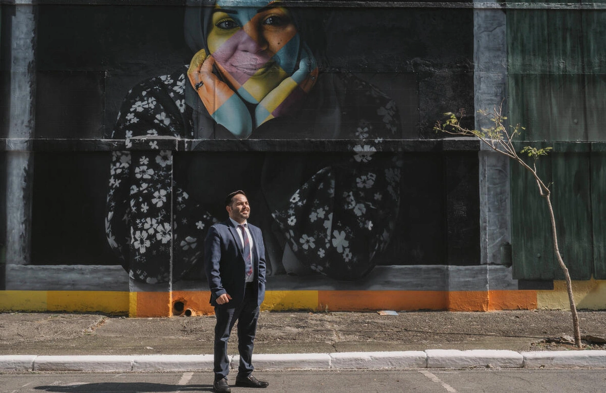 Man wearing a suit is standing in front of a wall with a huge drawing of a woman with a headscarf on it.