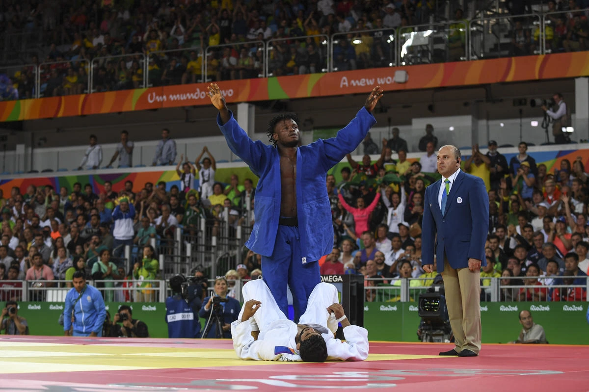 Un homme portant un uniforme bleu se tient debout, les mains en l'air en signe de victoire, tandis qu'un autre homme en blanc se couche en signe de défaite. Une foule nombreuse les entoure.