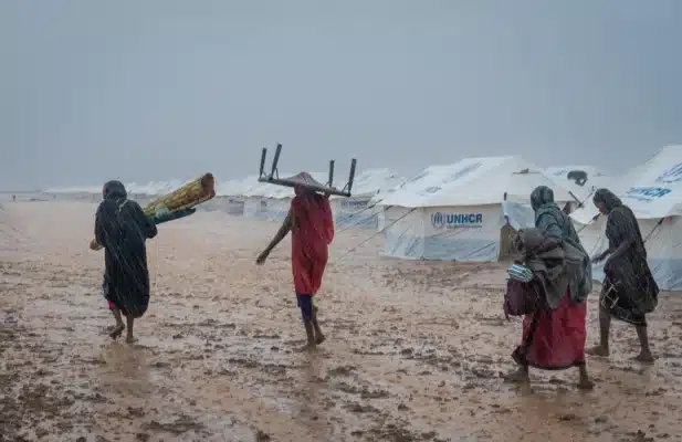 Four people are walking on a muddy ground under heavy rain as they are carry valuables. There are multiple UNHCR-labelled tents around them.