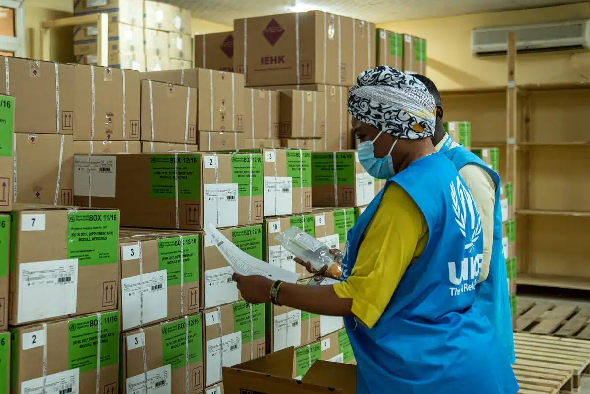 Une femme portant un masque chirurgical et un gilet bleu avec le logo du HCR en blanc examine des rangées de boîtes vertes dans un entrepôt.
