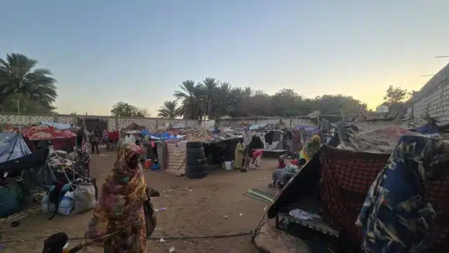 A woman dressed in a colourful dress and matching shawl covering her head leads a small child by hand on a sandy path among makeshift tents made of various materials as other women and children mill around. In the background, palm trees and a sunset are visible.