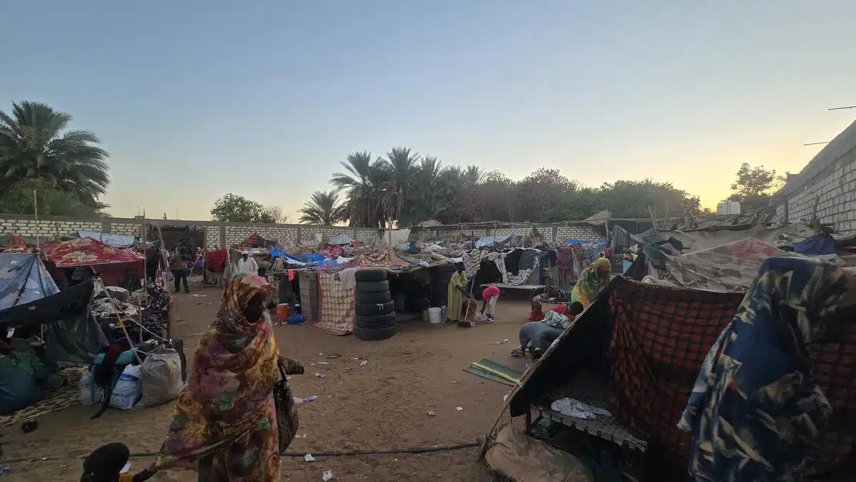 A woman dressed in a colourful dress and matching shawl covering her head leads a small child by hand on a sandy path among makeshift tents made of various materials as other women and children mill around. In the background, palm trees and a sunset are visible.