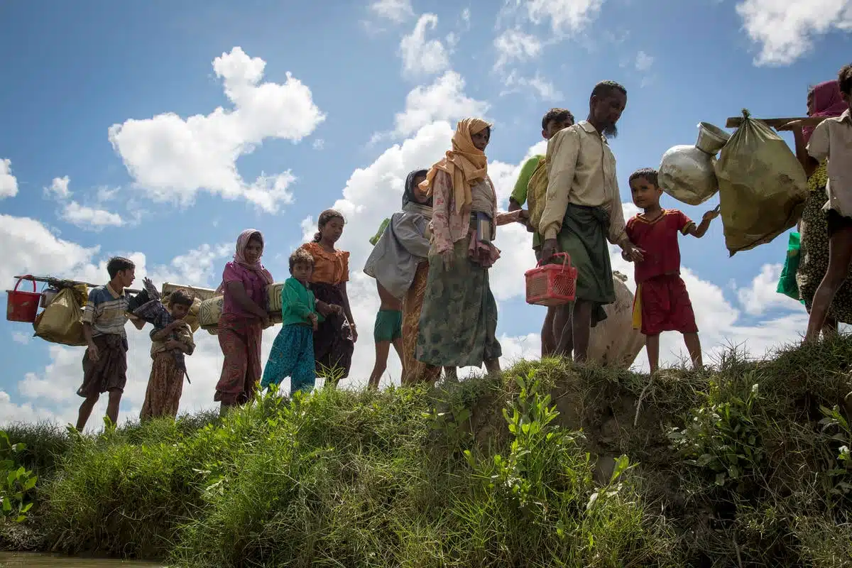 A group of Rohingya refugees, mostly women and children, carrying their belongings walk on a grassy knoll with bright blue sky and white clouds behind them in the background.