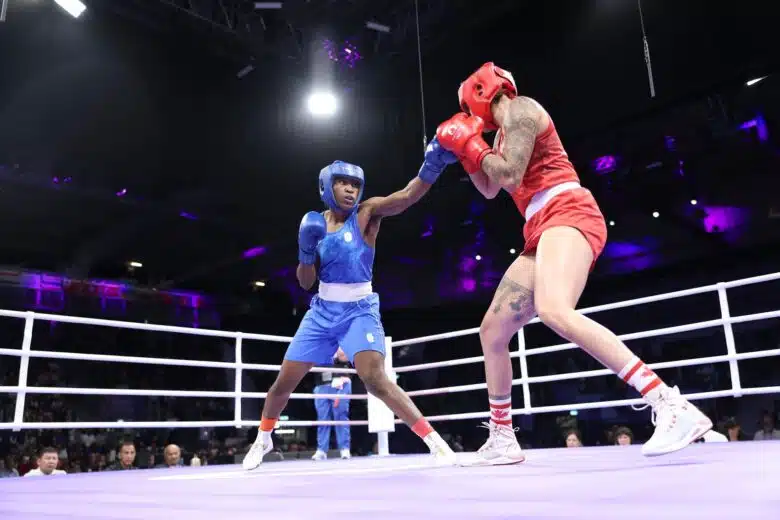 Two women in boxing ring. Woman on the left is in blue tank top and shorts with blue helmet and boxing gloves. She has her left arm extended. Woman on the right is in red tank top and shorts with red helmet and boxing gloves.