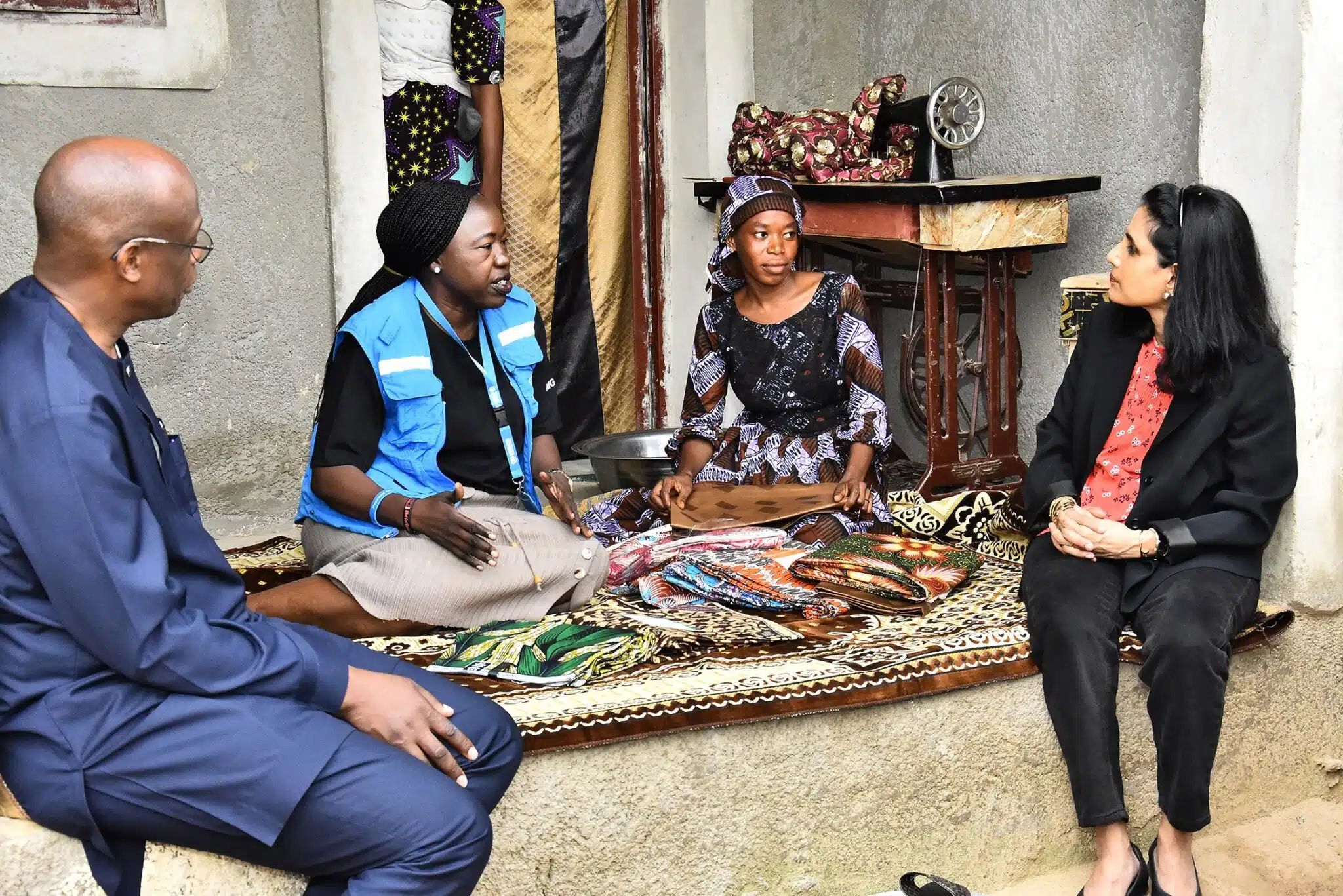 One man and three women talk while sitting on a raised floor covered with a carpet and fabrics. A young African woman dressed in brown and white dress and matching headdress sits next to an old pedal-powered sewing machine and shows pieces of colourful fabric to a South Asian woman dressed in a black pantsuit and an African woman in a blue UNHCR vest, while a black man dressed in a blue African style suit sits on the left.