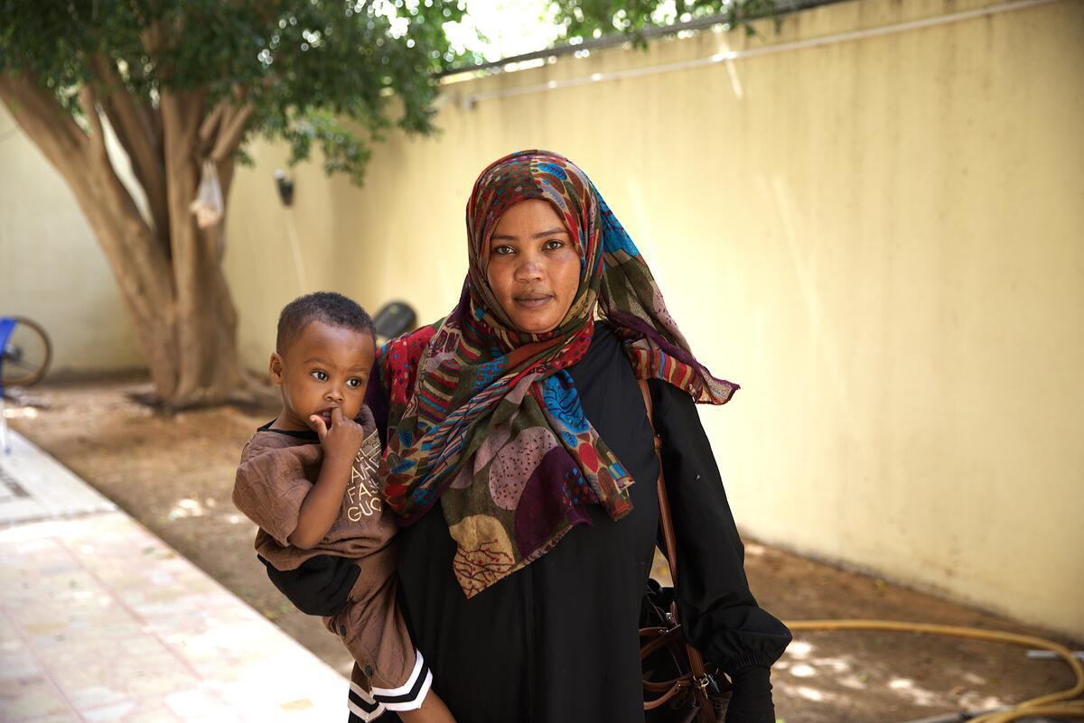 Person wears a dark-toned yet colourful head covering in addition to a black long sleeve shirt or dress. The centered person also is sporting a bag and carrying a child on their hip. They stand in a fenced in area with a bike wheel and tree in the background.
