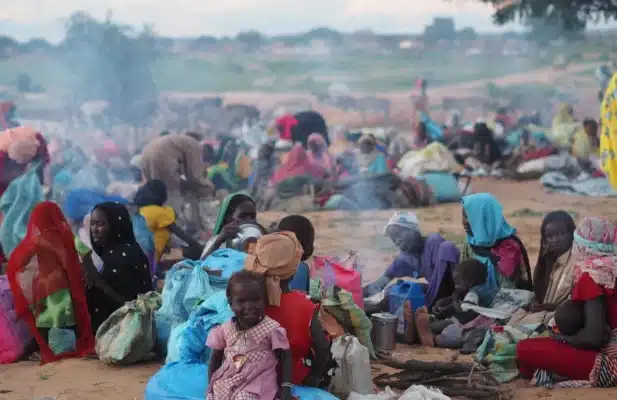 Families sit gathered around fires and pots upon a landscape of dirt and sparse grass.