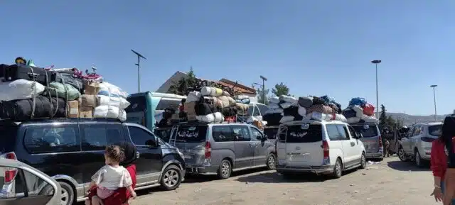 Several vans crowd a dirt roadway with luggage piled on top of the vehicles. They all are heading the same direction.