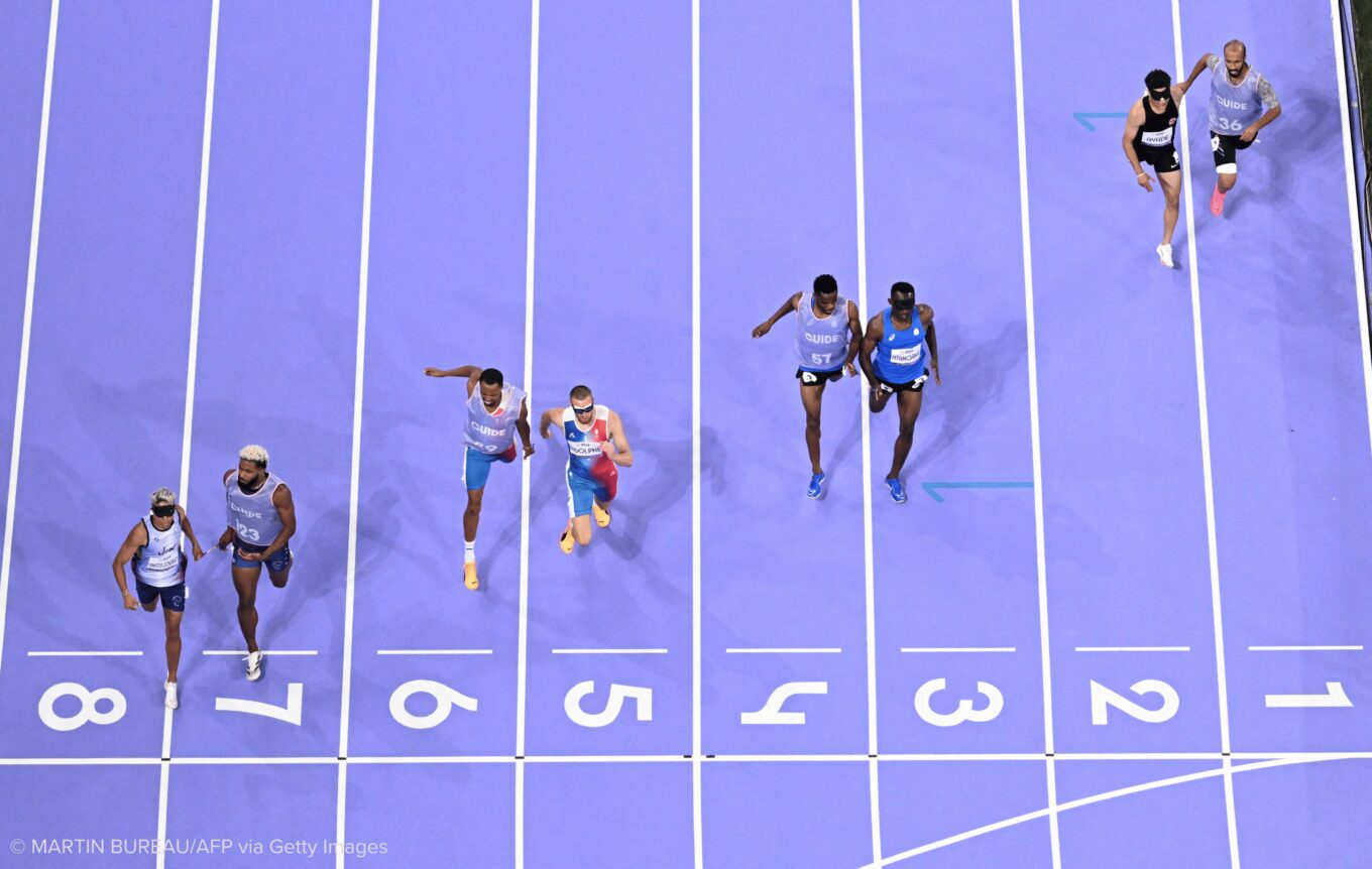Four athletes each paired with a guide person sprint towards a finish line. The athlete on the far left is closest to the finish line, each subsequent athlete descends in place, with the far right athlete as the very last of the bunch.