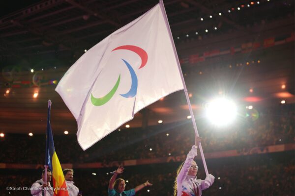 A young woman with a missing left forearm, wearing the white uniform of the Refugee Paralympic Team proudly carries a white flag with the symbol of Paralympic movement with red, blue, green asymmetrical crescents in the centre of the flag.