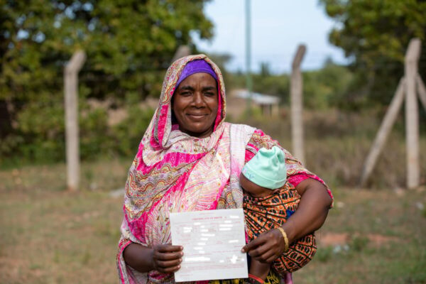 Woman proudly displays a birth certificate with a small child in her arms. The both of them wear very colourful and patterned clothing while standing in front of a fence with tall posts and greenery surrounding the two of them.