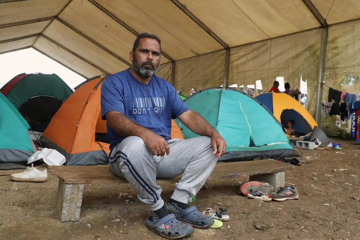 Man sits on a wooden bench with his hands resting on his knees. Behind him are three brightly coloured tents.