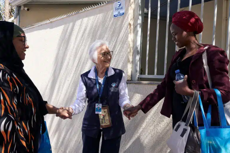 A woman stands in the middle of two women, smiling at the woman on her left as she takes one hand in each of hers.