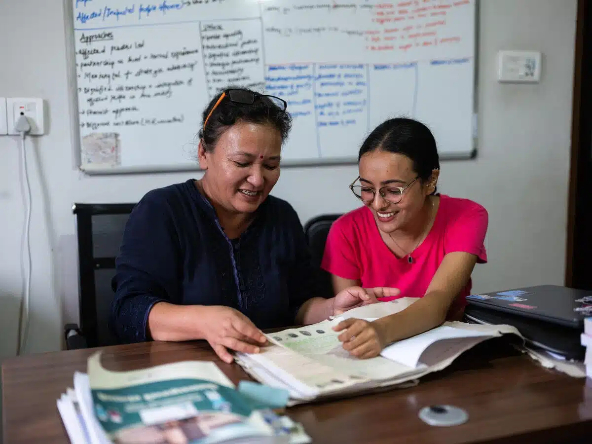 Deux femmes sourient alors qu'elles examinent des documents devant un tableau blanc sur lequel sont inscrites des informations.