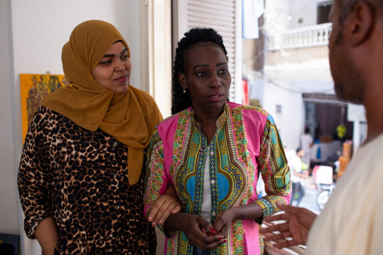 A woman holds her hands together with a serious expression on her face as she speaks to a man beside the camera. A different woman stands next to her and looks at the woman with her hands held together in front of her.