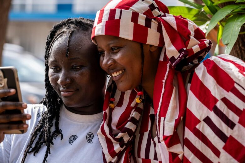 Two women pose for a photo. The one on the right wears a striped red and white outfit anf is displaying a broad smile. The woman on the left is smiling lightly while holding a phone.