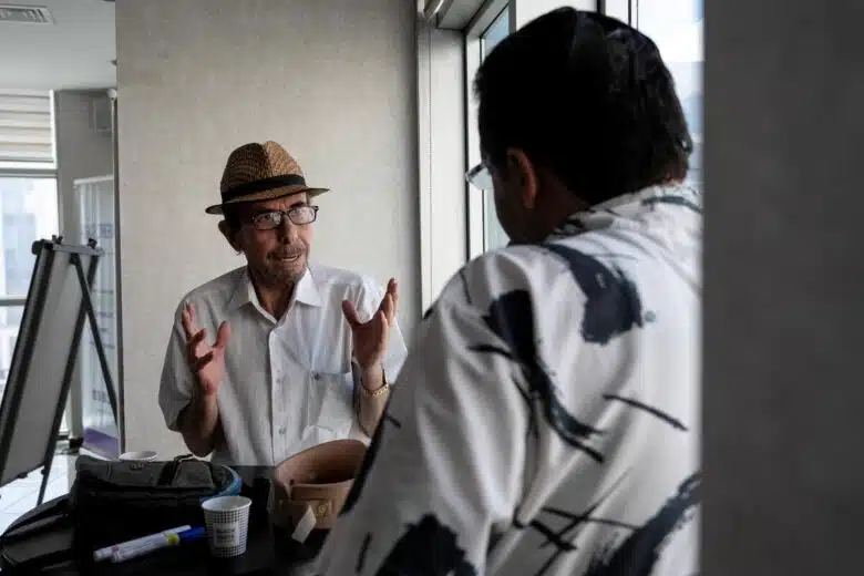 Deux hommes discutent. La caméra se concentre sur l'un d'entre eux qui parle de manière expressive avec ses mains, tandis que l'autre semble écouter, dos à la caméra.