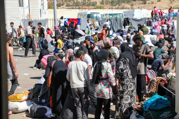 Families stand and sit on a dirt paved road with many belonings. Vehicles and humanitarian aid workers appear in the background and blankets are hung to dry on lines.