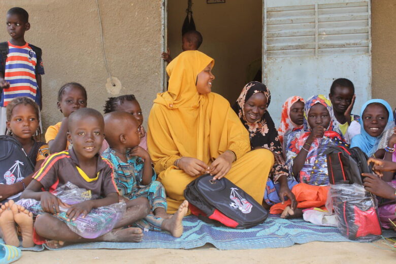 A woman wearing a bright yellow outfit sits on a blanket with several children in front of a door. She holds a backpack and smiles to children as they hold backpacks as well.