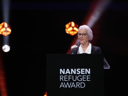 An elder woman speaks into a microphone attached to a podium. The background is black and filled with the occassional strobe light as professional lighting illuminates the podium in which she speaks from.