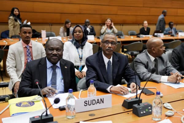 People sit lined behind a panel which displays "SUDAN".