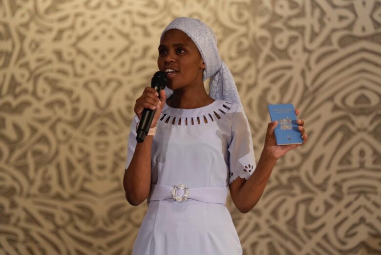 Woman stands in front of a wall adorned with patterned wallpaper. HSe speaks into a mic and holds a Kenyan passport in her opposite hand, presenting it. She wears a light purple dress and a matching headcovering.