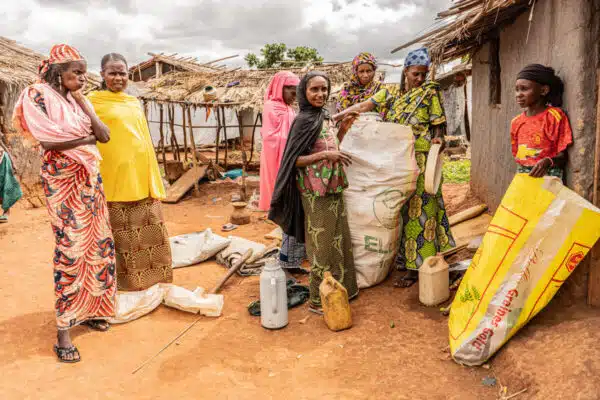 Deux femmes à gauche sont légèrement séparées du groupe de droite. Elles portent de longues robes culturelles colorées. Quatre femmes sur la droite, vêtues de vêtements culturels colorés, mettent des affaires dans un sac. Un jeune garçon se tient à leur droite et regarde à travers l'écran en tenant un sac jaune. Ils se tiennent sur une parcelle de terre et on peut voir des abris autour d'eux.