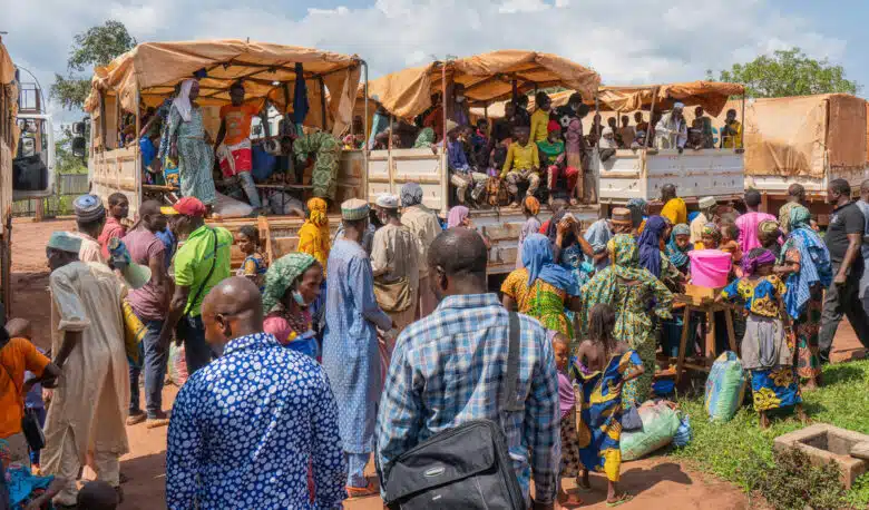 Quatre grands camions caravanes sont garés, l'arrière tourné vers la caméra. Les trois au centre sont remplis de plusieurs personnes portant des vêtements colorés. D'autres personnes se tiennent avec leurs familles au fond de l'arrière des camions.