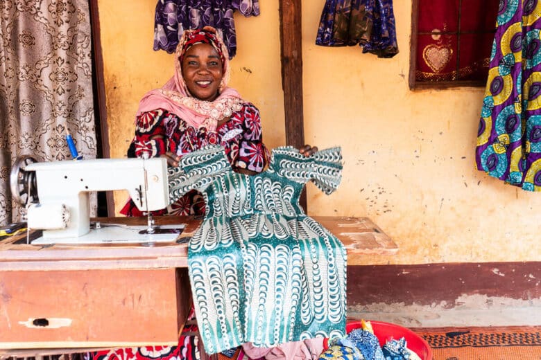 A person proudly displays a green and white dress while sitting in front of a yellow building and next to a sewing machine.Three other articles of clothing hang in the background.