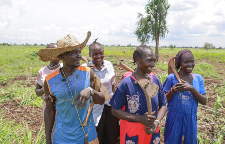 Five people smile toward the right side of the screen while holding farming tools.