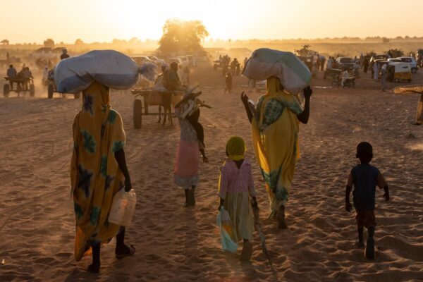 The camera is focused on five people, two adults and three children. Their backs are facing the camera as the carry supplies in the direction of the sun. The area is still darker than expected because of the sand's dust.