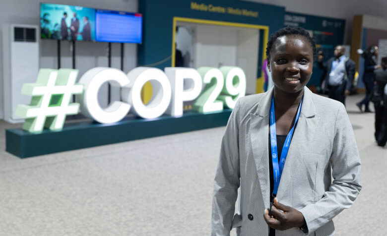 A person stands in front of a large display of "#COP29" which is dimly lit by green and yellow hues of light. The person is smiling and getnyl tugging their blazer closed.