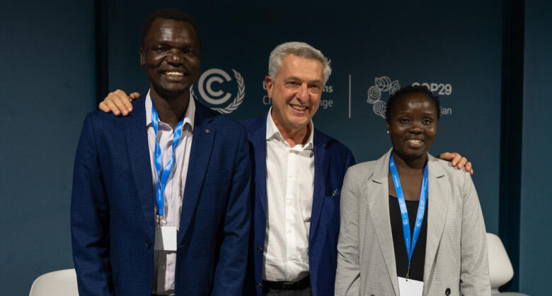 Three people stand left to right in order of tallest to least tall. The person on the middle joins the two on the sides by displaying a broad smile and placing a hand on each of their shoulders.