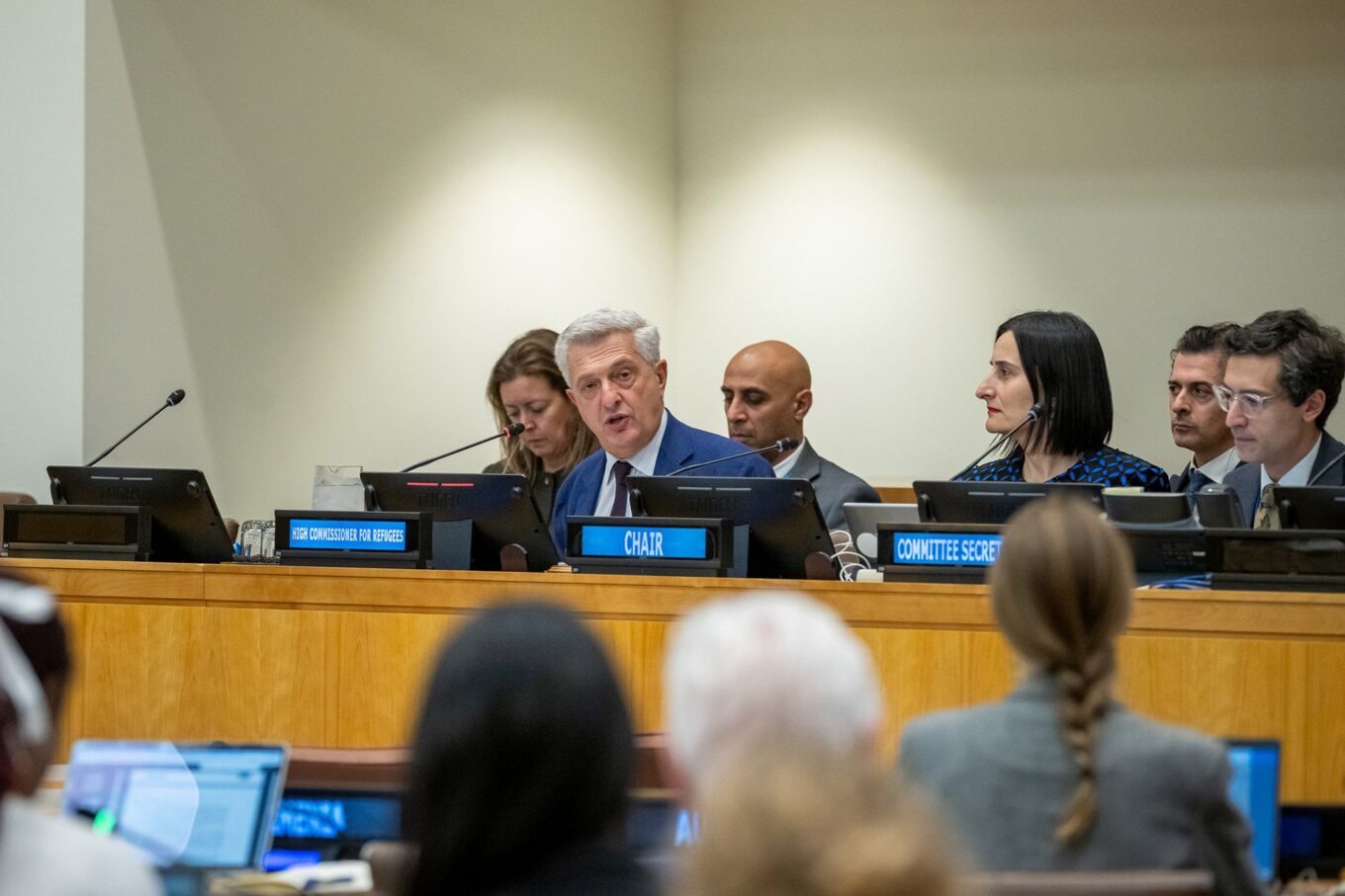 Three people sit behind three people who sit at the front of a raised and wooden speaker's panel. The first row features name plates that display the following (left to right): High Commissioner for Refugees, Chair and Committee Secretary. The person sitting behind the first name plate appears to be speaking. People sit below and observe the discussion.